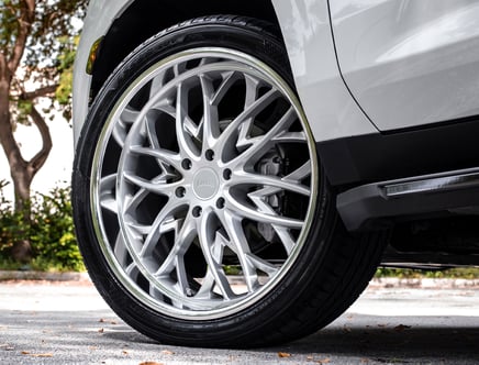 Close-up of a stylish multi-spoke alloy wheel on a gray vehicle, set against a leafy outdoor background.