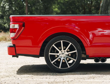 Rear side view of a red pickup truck, focusing on the wheel, part of the bed, and dual exhaust pipes, parked on a gravel surface with trees in the background.