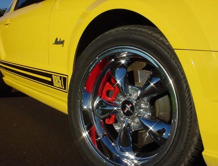 Close-up of a yellow car's front wheel, featuring shiny chrome rims, red brake calipers, and "Mach 1" text on a black stripe along the side of the vehicle.