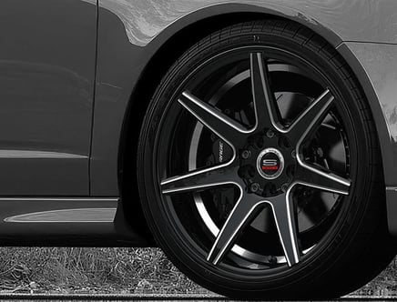 Close-up of a car's black alloy wheel with a silver outer rim and a central hubcap displaying the letter 'S' beneath a silver vehicle body.