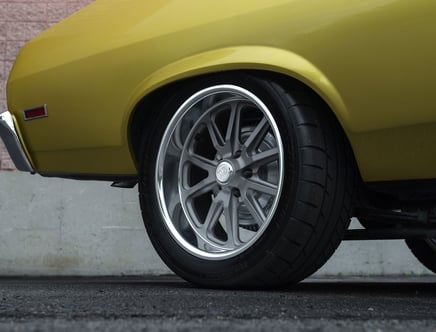 Close-up of a yellow car's rear wheel featuring a silver alloy rim and a low-profile tire on a textured road surface.