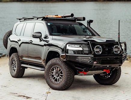 A black off-road SUV equipped with large tires, a rooftop rack, and specialized front bumper lights, parked near a body of water.