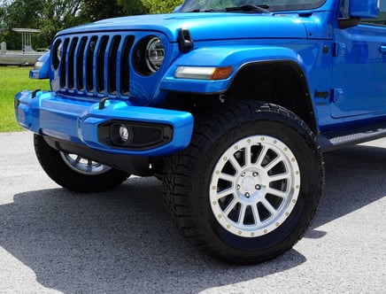 Close-up view of the front half of a blue Jeep Wrangler, highlighting its large tires and distinctive grille on a sunny day.