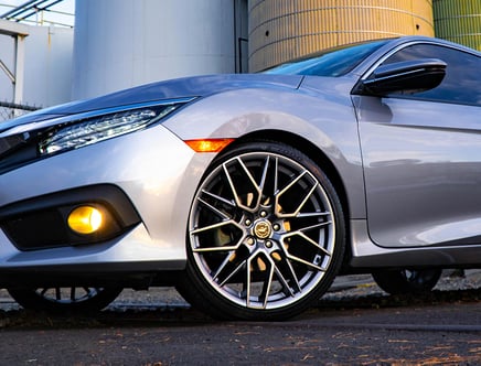 Low angle view of the front corner of a silver car with distinctive alloy wheels and illuminated headlights, parked on a street with industrial buildings in the background.