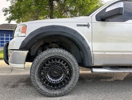 Close-up of the front side of a silver pickup truck with black fender flares and a large black GFX tire. The truck is parked on a street with a green tree in the background.