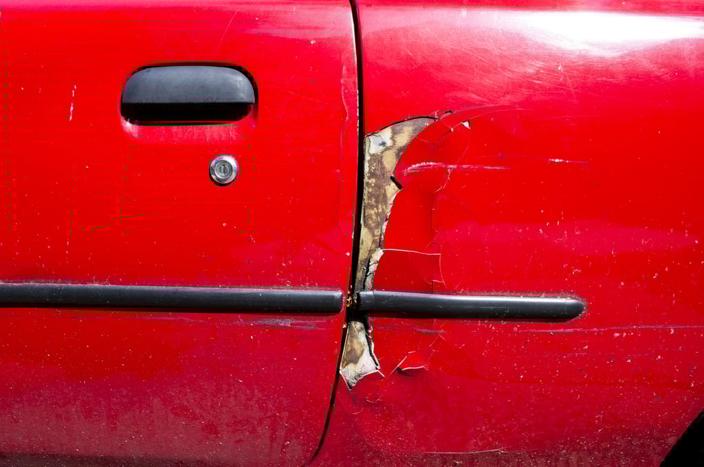 Closeup of a red car with old and large paint chips showing early stages of rusting and corrosion.