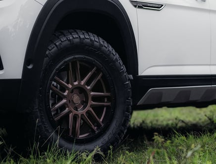 Close-up view of a vehicle's rear wheel and tire on grassy terrain, featuring a rugged, multi-spoke alloy rim and visible tire tread pattern.