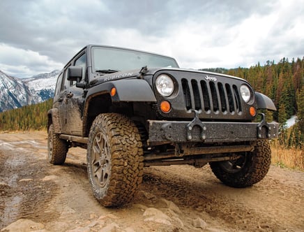 A muddy black Jeep Wrangler Rubicon on a dirt mountain trail with snow-capped peaks and a forest background.