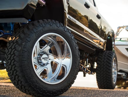 Close-up of a lifted black truck with oversized Moto Metal wheels and aggressive tread tires, parked near a boat and water in the background.