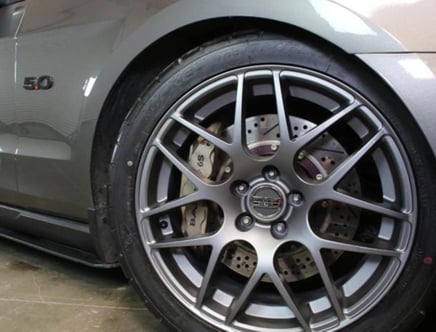 Close-up of a car's wheel featuring a multi-spoke alloy rim, branded with "Forgestar," and visible brake components behind it, with part of the gray car body and a "5.0" badge in the background.