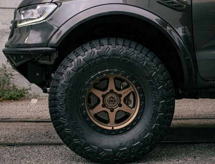 Close-up of a rugged off-road vehicle tire with bold, deep treads and a bronze-colored wheel rim, parked on a street.