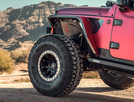 Close-up of the rear wheel and body of a red off-road vehicle, featuring large, rugged tires branded with "Patagonia Milestar" in a desert landscape.