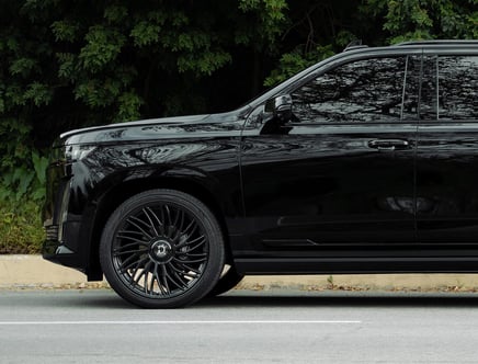 Side view of a sleek black SUV with large, stylish black wheels, parked on a street with green foliage in the background.