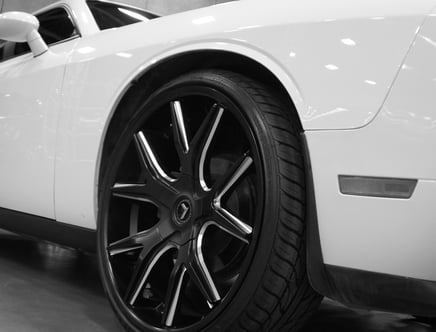 Black and white image of a white car featuring a close-up of its front tire and alloy wheel.