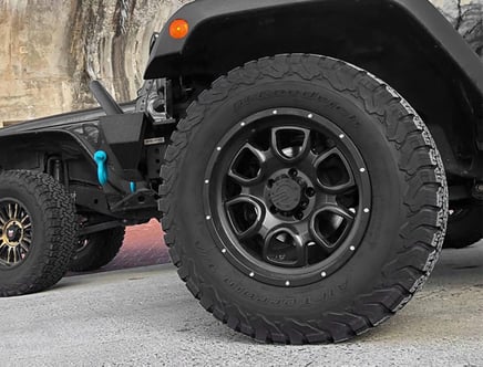 Close-up of a black vehicle tire with rugged tread, mounted on a black alloy wheel, next to another vehicle with similar off-road tires, parked on a concrete surface.