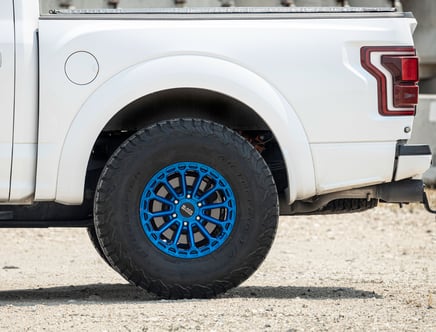 The rear section of a white pickup truck featuring a distinctive blue rim and all-terrain tire parked on a gravel surface.