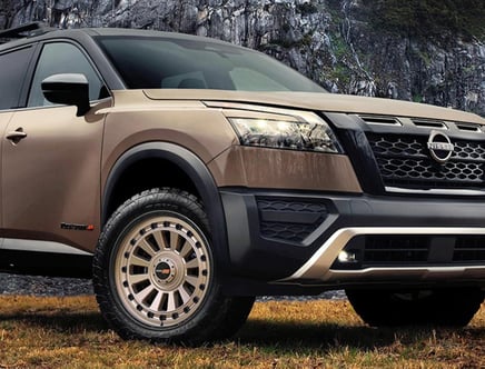 A close-up view of the front side of a brown Nissan SUV with rugged off-road tires, parked on grassy terrain against a rocky cliff background.