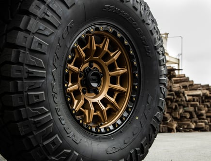 Close-up of a rugged off-road tire with a bronze custom wheel and distinct tread pattern, labeled "Ridge Grappler," against a backdrop of stacked wooden pallets.