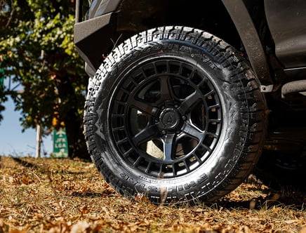 Close-up of a black off-road tire with a rugged tread pattern on a vehicle parked on dry grass with fallen leaves, with a tree in the background.