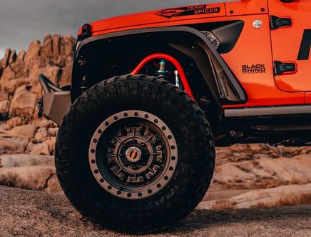 Close-up of an off-road vehicle's large, rugged tire set against a rocky landscape, featuring an orange body with "Black Rhino Off Road Wheels" branding.