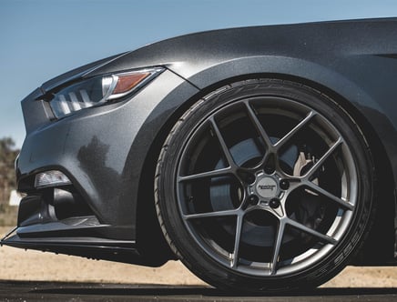 Close-up of the front wheel and headlight of a dark-colored sports car with American Racing wheels, under a clear blue sky.