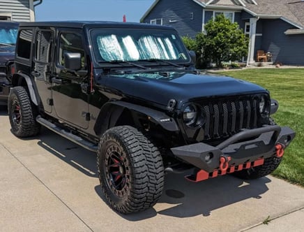 A black off-road vehicle parked on a driveway, equipped with large tires, red towing hooks, and a reflective sunshade on the windshield; a house and a lawn are visible in the background.