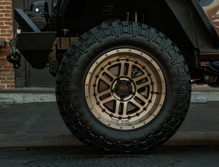 Close-up of an off-road vehicle tire featuring a black and beige "Moto Metal" wheel with a rugged tread pattern, parked on a dark surface with a brick wall background.