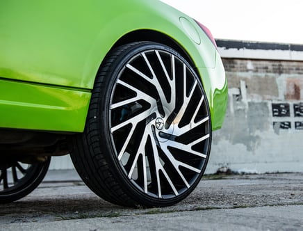 Close-up view of a lime green car with large custom black and silver rims, parked on a street with a weathered brick wall in the background.