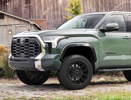A green Toyota Tundra pickup truck parked in front of a rustic wooden building, featuring a prominent hexagonal grille and large off-road tires.