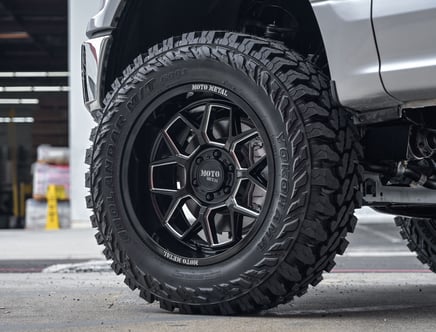 Close-up of a rugged tire on a vehicle, featuring black Moto Metal alloy wheels and a Yokohama Geolandar M/T G003 tire, set against a concrete garage floor.