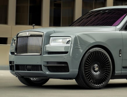 Front view of a luxurious silver car with a prominent grille, a sleek front bumper, and large black alloy wheels, parked in an urban setting.