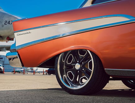 Close-up of the rear section of a classic Chevrolet car with an orange and white exterior parked on a tarmac beside an airplane, with a focus on its custom black and chrome wheel rims.