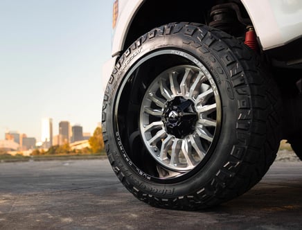 Close-up of a vehicle's off-road tire with a city skyline in the background, featuring a large, rugged tread pattern and a custom alloy wheel.