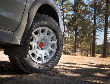 Close-up of a car's rear wheel with Yokohama Geolandar A/T tires parked on dirt in a forest setting.