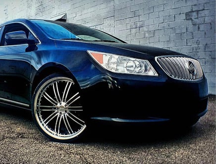 A black car with chrome rims parked next to a brick wall, displaying a distinctive grille and headlight design.