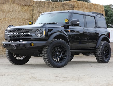 A black Ford Bronco with lifted suspension and large off-road tires, parked on a dirt surface in front of stacked hay bales.