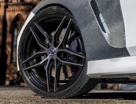 Close-up of a black alloy wheel with a distinctive spoke design on a white car, highlighting a carbon fiber fender and a slotted brake rotor behind the wheel.