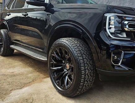 Close-up of a black SUV with large, rugged tires and sleek alloy wheels, parked on a dirt ground beside a gray wall.