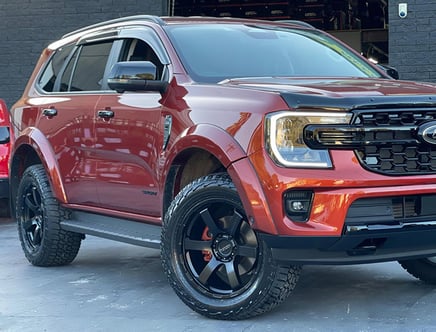 A red SUV with black wheels and a sleek, modern design is parked on a concrete surface. It has a distinctive grille and headlight design, and the vehicle appears to be polished and well-maintained, reflecting light on its surface.