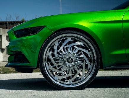Close-up of a green car with shiny chrome alloy wheels featuring an intricate spiral design, parked on a concrete surface.