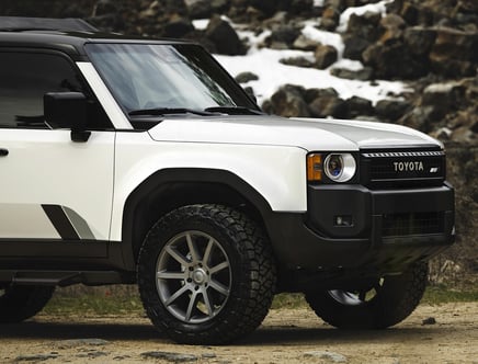 Partial view of a white Toyota SUV with distinctive black trim, parked on a dirt ground with rocky terrain in the background.