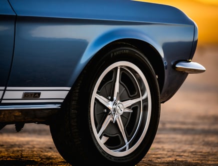 Close-up of a blue classic car's rear wheel and fender, featuring a GTA badge and chrome accents in a sunset setting.
