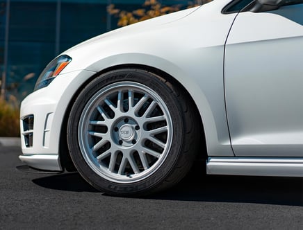 Close-up of a white car's front fender and wheel with a silver multi-spoke rim and low-profile Falken Azenis tire, parked on a paved surface.