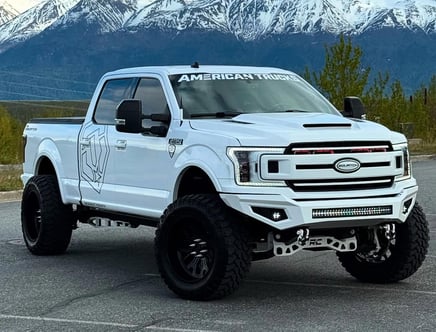 A customized white pickup truck with a prominent logo on the side parked in front of snow-capped mountains.