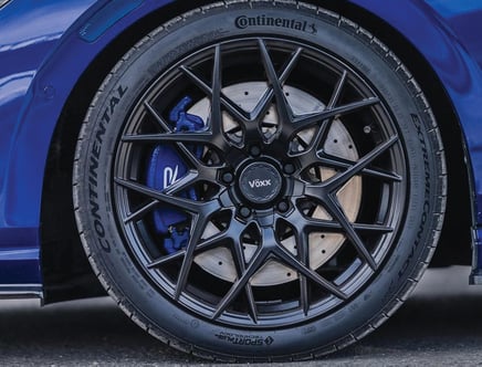 A close-up view of a car wheel featuring a black multi-spoke Voxx rim and a Continental tire. The blue brake caliper is visible behind the rim, and the tire model is ExtremeContact. The car's blue bodywork is partially visible.