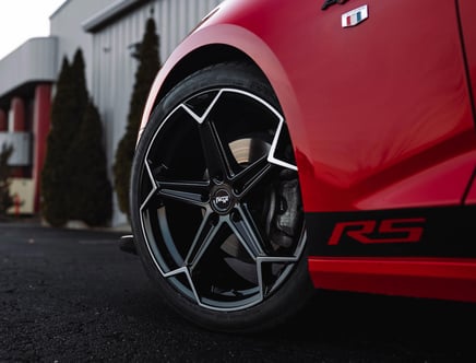 Close-up of a red sports car's front wheel featuring black and silver Niche alloy rims with an RS badge on the side, parked in front of a building with tall bushes.