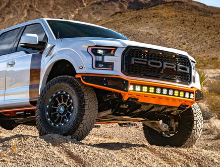 A white Ford truck with off-road tires and an orange front bumper, equipped with a row of LED lights, parked in a desert landscape with rocky terrain and mountains in the background.