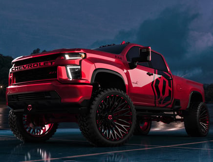 A modified red Chevrolet pickup truck with custom wheels and lifted suspension parked on a wet surface under a dusk sky.