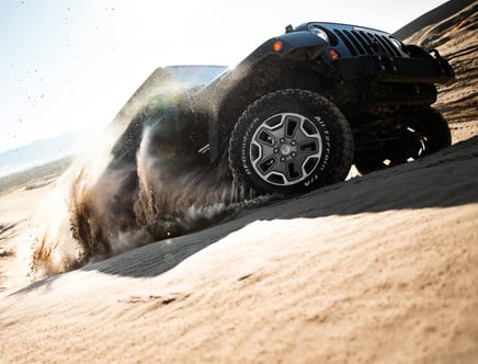 Off-road vehicle driving through sand dunes with tires kicking up sand.