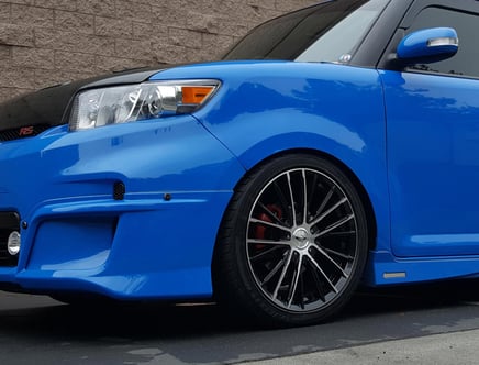 A blue car with custom black alloy wheels and a modified front bumper, parked in front of a brick wall.